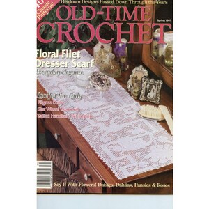May include: The cover of "Old-Time Crochet" magazine, Spring 1997, features a white floral filet dresser scarf. The magazine's title is in large, pink letters. Other items on the dresser include a brush, framed photos, and decorative bottles.