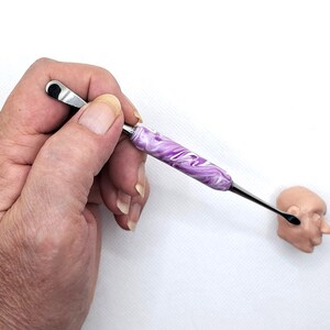May include: A tool with a stainless steel blade and a purple and white marbled handle. The tool is held near a small, light-brown sculpture of a human head. The tool has a small, flat blade on one end and a curved blade on the other.