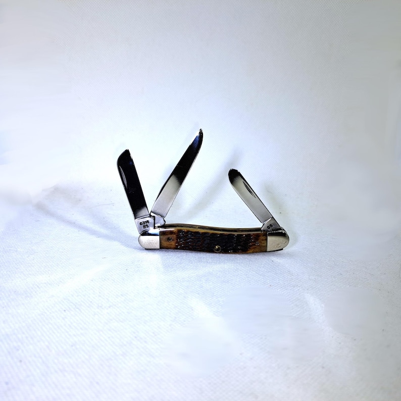 May include: A pocket knife with three open blades. The knife has a brown handle with a textured pattern and silver-colored metal accents. The blades are shiny and reflect the light. The knife is set against a white background.
