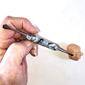 May include: A silver-colored ear cleaning tool with a marbled black and white handle. The tool is held near a small, tan-colored plastic head.