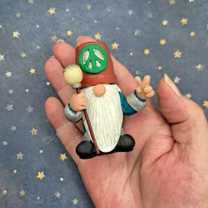 May include: A small gnome figurine with a white beard and a red hat with a green peace sign. The gnome is holding a wooden staff with a white ball on top. The gnome is standing on a hand.