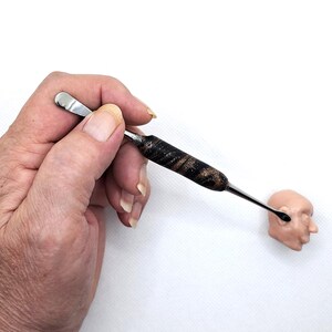 May include: A hand holds a black and gold sculpting tool with a metal tip. The tool is being used to sculpt a small, light brown clay head.
