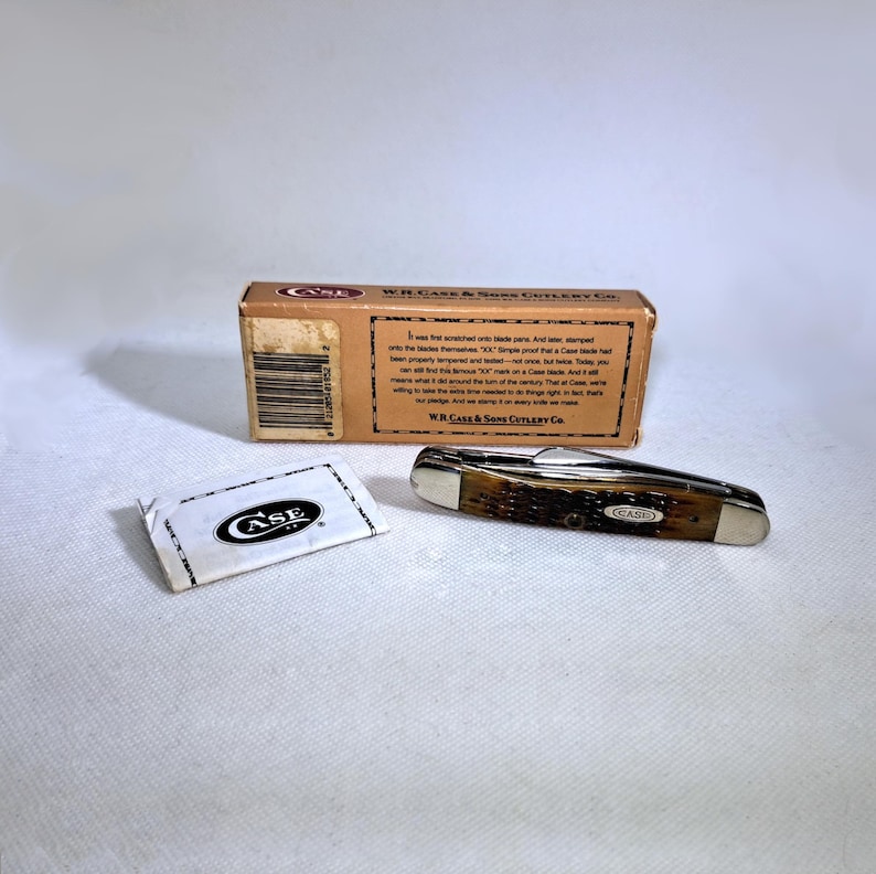 May include: A Case pocket knife with a brown handle and silver accents, next to its original box and a small packet with the Case logo. The box has text and a barcode. The knife is closed, showing the blade's edge.