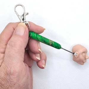 May include: A green and gold marbled tool with a silver metal tip is held by a silver clip. The tool is positioned near a small, pale-skinned sculpture of a human head.