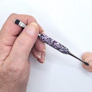 May include: A silver and black tool with a patterned purple and white handle is held near a small, light-colored head-shaped object. The tool has a small, flat, black tip on one end and a rounded black tip on the other.
