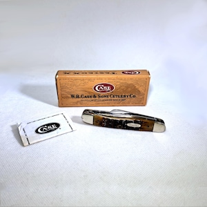 May include: A Case pocket knife with a brown handle and silver accents, next to its original box and a small packet. The box reads "W.R. Case & Sons Cutlery Co." and "Hand-Crafted Knives Since 1889."