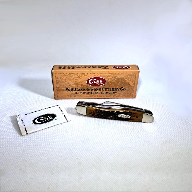 May include: A Case pocket knife with a brown handle and silver accents, next to its original box and a small packet. The box reads "W.R. Case & Sons Cutlery Co." and "Hand-Crafted Knives Since 1889."