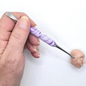 May include: A hand holds a purple and white marbled sculpting tool with a metal tip. The tool is being used to sculpt a small, unpainted clay head.