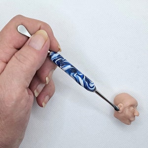 May include: A sculpting tool with a stainless steel blade and a handle with a blue and white swirl pattern. The tool is being used on a small, tan clay head. The tool is approximately 6 inches long.