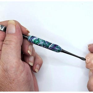 May include: A double-ended sculpting tool featuring a central handle with a marbled pattern of green, purple, and white swirls. The tool has a flat, rounded metal tip on one end and a smaller, curved metal tip on the other, used for shaping clay.