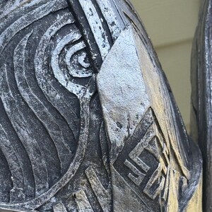 May include: Close-up of a metallic, silver-toned object with intricate, embossed patterns. The design features swirling lines, geometric shapes, and a diamond-shaped emblem with stylized symbols. The object has a weathered appearance.