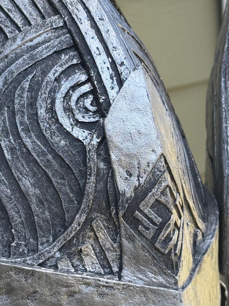May include: Close-up of a metallic, silver-toned object with intricate, embossed patterns. The design features swirling lines, geometric shapes, and a diamond-shaped emblem with stylized symbols. The object has a weathered appearance.