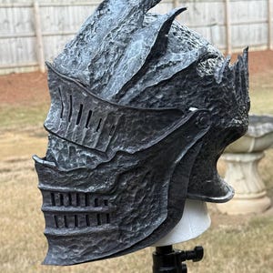 Puede incluir: Un casco gris oscuro texturizado con visera y parte superior puntiaguda. El casco tiene un diseño medieval, con ranuras verticales en la visera y una superficie texturizada. El casco está montado sobre un soporte blanco.