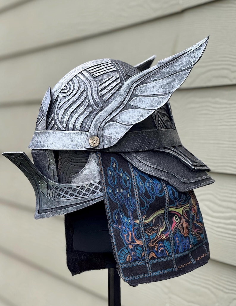 May include: A silver-colored helmet with wing-like accents and intricate carvings. The helmet has a decorative button and is paired with a black and blue patterned fabric. The design suggests a fantasy or historical theme.