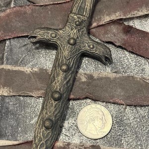 Może przedstawiać: Szczegółowy sztylet w stylu antycznym z teksturowanym, metalicznym wykończeniem. Sztylet ma rękojeść w kształcie krzyża i szereg okrągłych zdobień wzdłuż rękojeści. Moneta jest umieszczona obok sztyletu w celu porównania rozmiaru.
