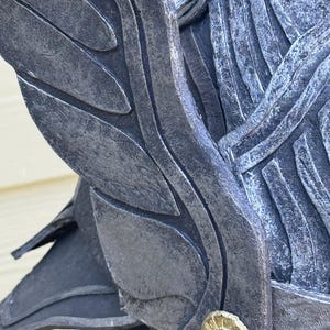 May include: Close-up of a dark gray helmet with wing-like details. The helmet has a textured, weathered appearance with a gold-colored circular emblem. The design suggests a fantasy or historical theme.