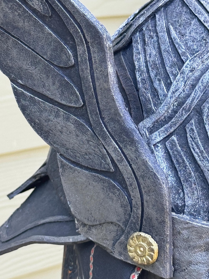 May include: Close-up of a dark gray helmet with wing-like details. The helmet has a textured, weathered appearance with a gold-colored circular emblem. The design suggests a fantasy or historical theme.