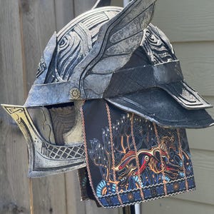May include: A silver and black fantasy helmet with wing-like accents and intricate detailing. The helmet features a decorative face guard and a black fabric neck guard with a colorful, patterned design. The helmet is displayed against a wooden background.