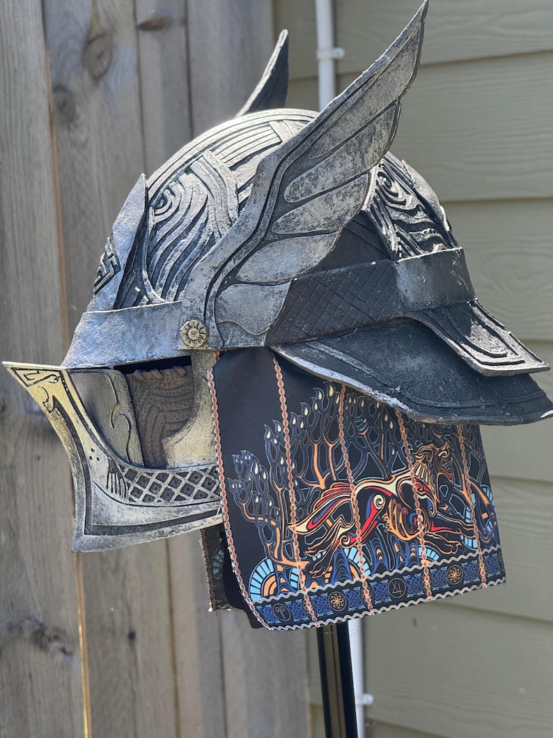 May include: A silver and black fantasy helmet with wing-like accents and intricate detailing. The helmet features a decorative face guard and a black fabric neck guard with a colorful, patterned design. The helmet is displayed against a wooden background.