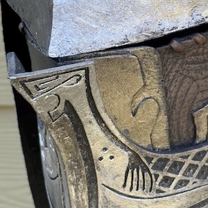 May include: Close-up of a metallic, possibly armor piece. The design features intricate carvings with a silver and gold color scheme. The details include a stylized hand and geometric patterns.