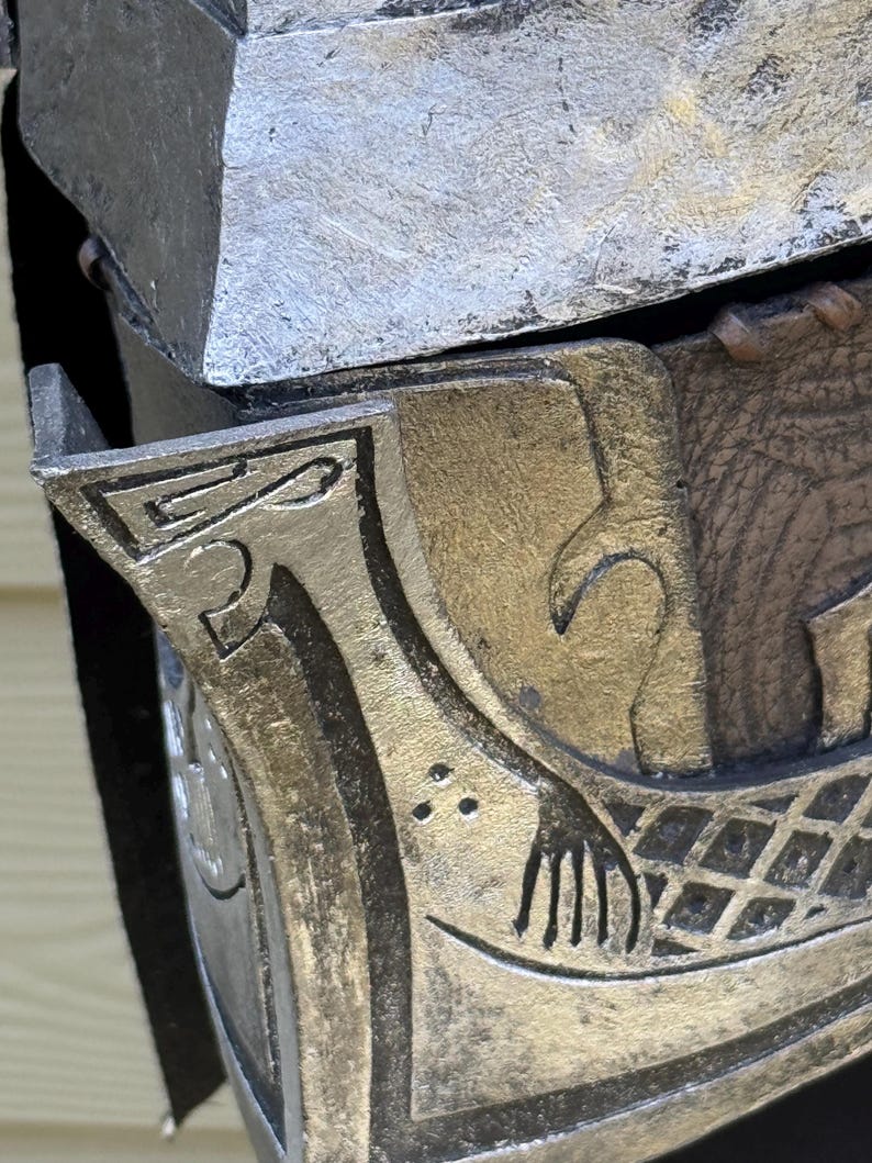 May include: Close-up of a metallic, possibly armor piece. The design features intricate carvings with a silver and gold color scheme. The details include a stylized hand and geometric patterns.