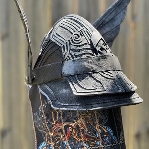 May include: A black helmet with silver accents and wing-like protrusions. The helmet features layered plates and an intricate design on the front and lower portion, with a colorful, patterned fabric. The helmet is likely for costume or display.