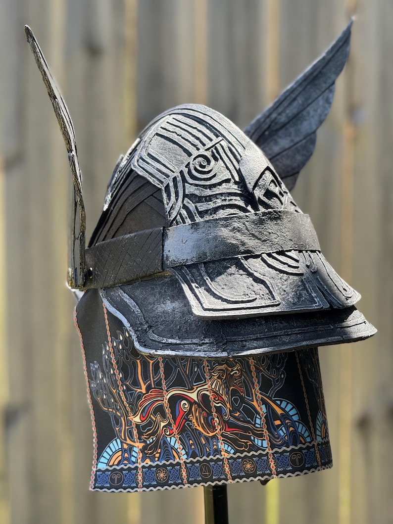 May include: A black helmet with silver accents and wing-like protrusions. The helmet features layered plates and an intricate design on the front and lower portion, with a colorful, patterned fabric. The helmet is likely for costume or display.