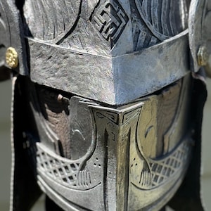 May include: A detailed close-up of a silver-colored helmet with intricate carvings. The helmet features a prominent crest, geometric patterns, and a visor with a boat-like design. The metallic surface has a weathered appearance.