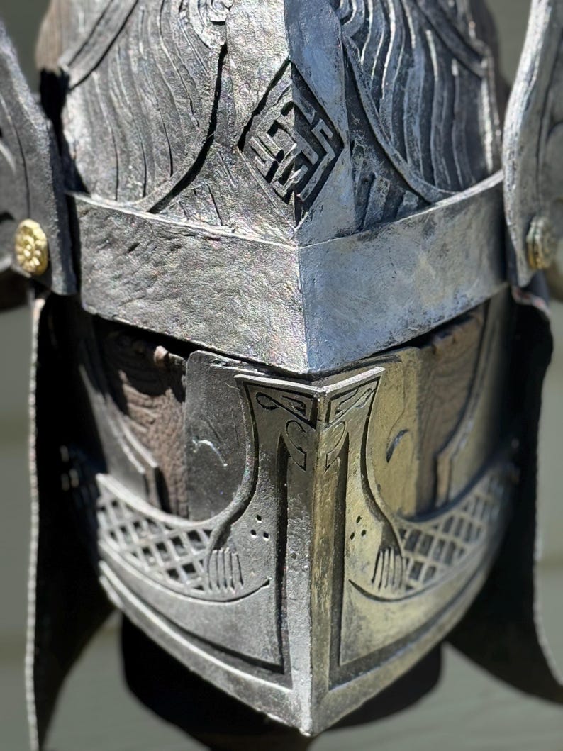 May include: A detailed close-up of a silver-colored helmet with intricate carvings. The helmet features a prominent crest, geometric patterns, and a visor with a boat-like design. The metallic surface has a weathered appearance.