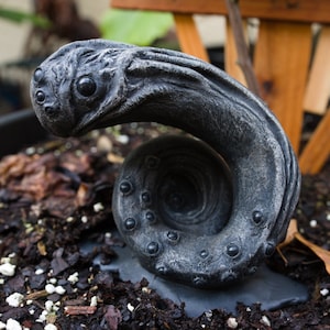 May include: A gray, textured, curled, and twisted garden statue with multiple eyes and a circular opening in the center.