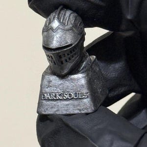 May include: A small, silver-colored figurine of a knight's helmet and shoulders. The figurine has a weathered appearance and the words "DARK SOULS" are inscribed on the base. The knight's helmet has a visor with rectangular openings.