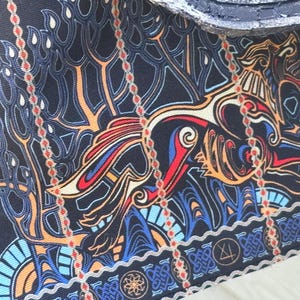 May include: Close-up of a fabric with a detailed, colorful design. The pattern features a stylized dragon or mythical creature in shades of red, blue, orange, and gold against a dark blue background. The design includes intricate lines and shapes.