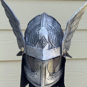 May include: A silver-colored helmet with intricate carvings and two large, stylized wings. The helmet features a pointed front and a decorative emblem. The wings are attached on either side, adding to the helmet's dramatic appearance.