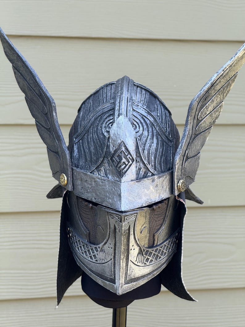 May include: A silver-colored helmet with intricate carvings and two large, stylized wings. The helmet features a pointed front and a decorative emblem. The wings are attached on either side, adding to the helmet's dramatic appearance.