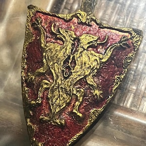 May include: A gold and red shield pendant featuring an intricate design of a double-headed wolf. The pendant has a loop at the top for hanging.