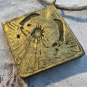 May include: A gold-colored square pendant with a raised design of a figure holding a crescent moon and stars. The pendant is attached to a brown cord.