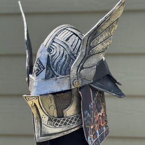 May include: A silver and black fantasy helmet with wing-like accents. The helmet features intricate detailing, including etched patterns and a decorative face guard. The design incorporates a mix of metallic and dark elements, suggesting a warrior or mythical theme.