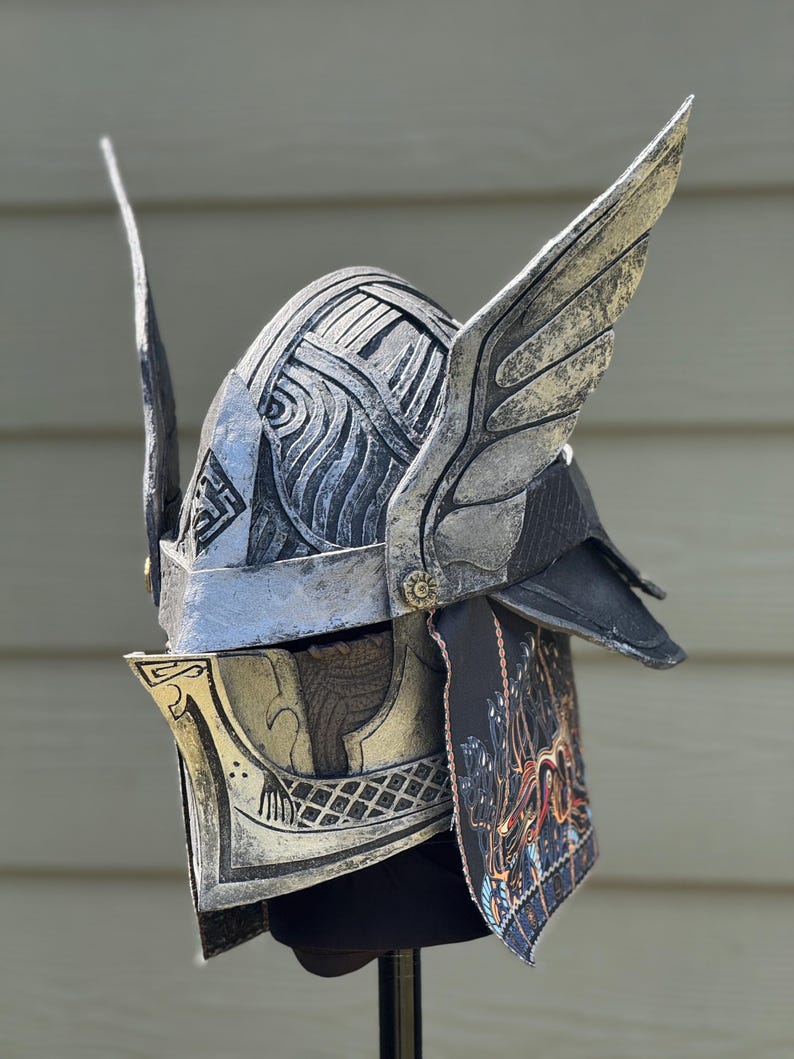 May include: A silver and black fantasy helmet with wing-like accents. The helmet features intricate detailing, including etched patterns and a decorative face guard. The design incorporates a mix of metallic and dark elements, suggesting a warrior or mythical theme.