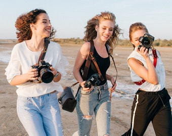 Könnte beinhalten: Drei Frauen in legerer Kleidung mit Kameras gehen an einem Sandstrand entlang. Sie lächeln und lachen, während sie gehen.