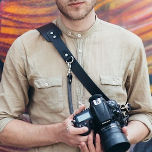 Può includere: Una persona che indossa una camicia abbottonata color beige chiaro con una tracolla per fotocamera in pelle nera e una fotocamera nera con un obiettivo argentato. La persona indossa occhiali da sole color blu e ha i capelli corti castani.