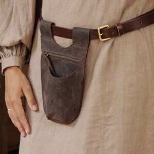 May include: A brown leather belt with a brown leather pouch attached. The pouch has a zipper and a small pocket. The belt is worn over a light brown linen dress.