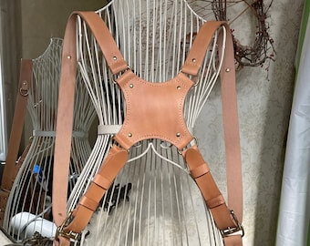 May include: A tan leather camera strap with adjustable straps and brass hardware. The strap is designed to be worn across the chest and back.