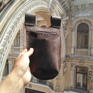 May include: A brown leather pouch with a zippered pocket and a flap closure. The pouch is held in a hand against a background of an ornate building.
