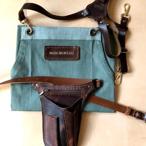 Puede incluir: Un delantal verde salvia con un parche de cuero marr&oacute;n con el texto MON BUREAU, combinado con correas de hombro de cuero marr&oacute;n oscuro y una funda de herramientas de cuero marr&oacute;n con bolsillo con cremallera y el texto ZAG.