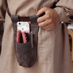 May include: A brown leather belt with a brown leather pouch attached. The pouch has a zipper and a flap that closes with a snap. A cell phone and two red tools are inside the pouch.