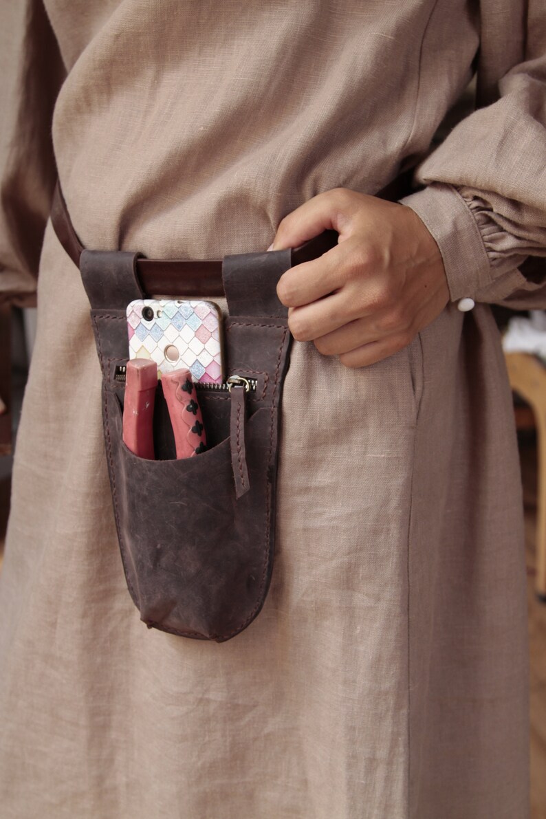 May include: A brown leather belt with a brown leather pouch attached. The pouch has a zipper and a flap that closes with a snap. A cell phone and two red tools are inside the pouch.