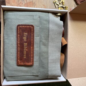 Puede incluir: Un delantal de lona verde salvia plegado dentro de una caja de cart&oacute;n. El delantal presenta un parche rectangular de cuero marr&oacute;n cosido en la parte delantera con el texto Ergo Bibamus grabado en oro. Un anillo de metal color lat&oacute;n est&aacute; unido a la esquina superior.