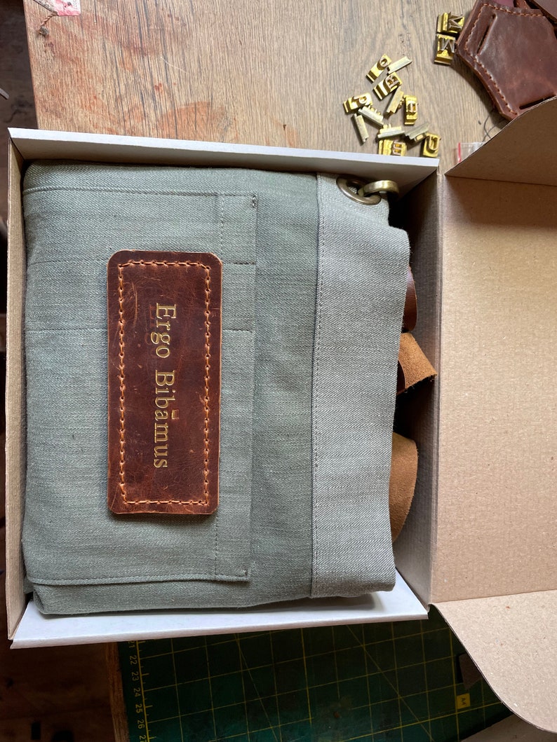 Puede incluir: Un delantal de lona verde salvia plegado dentro de una caja de cart&oacute;n. El delantal presenta un parche rectangular de cuero marr&oacute;n cosido en la parte delantera con el texto Ergo Bibamus grabado en oro. Un anillo de metal color lat&oacute;n est&aacute; unido a la esquina superior.