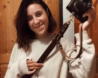 May include: A dark brown leather camera strap with gold-colored hardware is attached to a black camera. The strap is held by a person wearing a light-colored sweatshirt. Text on the image reads "Camera strap from @merifa_leather? Hell yeah Maybe I feel badass now".