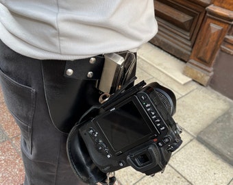Puede incluir: Una cámara negra con una funda de cuero unida a un cinturón negro. La cámara tiene una lente grande y una pantalla en la parte posterior.